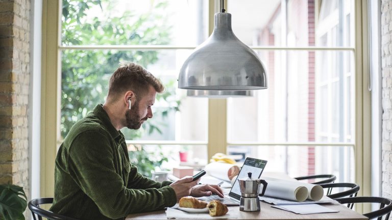 Eine Person sitzt an einem Esstisch vor einem Notebook und hält ein Smartphone in der Hand.