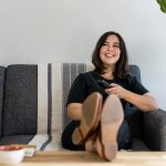 Eine Frau sitzt auf dem Sofa, legt die Beine entspannt auf dem Tisch ab und h&auml;lt in der Hand eine Fernbedienung.