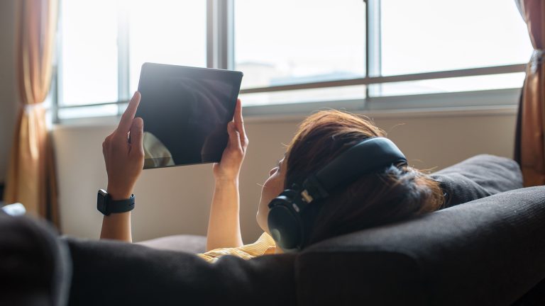 Eine Frau liegt auf dem Sofa, in der Hand ein Tablet, auf dem Kopf einen Kopfhörer.