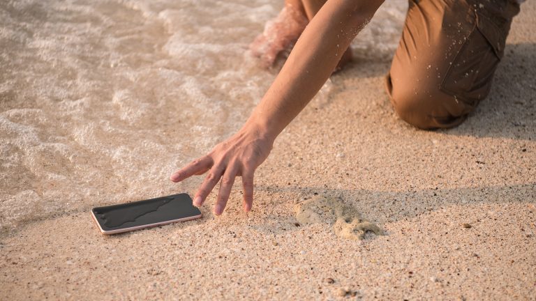 Ein Handy liegt am Strand und wird von Wasser umspült, eine Hand greift nach dem Gerät.