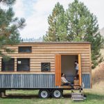 Zwei Personen sitzen am Eingang eines Tiny House, das auf einen Anh&auml;nger gebaut ist.