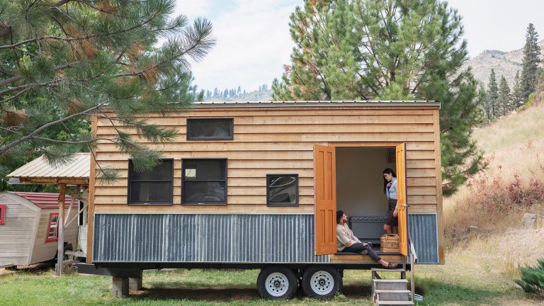 Zwei Personen sitzen am Eingang eines Tiny House, das auf einen Anh&auml;nger gebaut ist.
