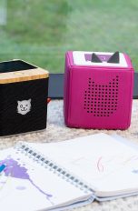 Eine schwarze Tigerbox Touch steht neben einer pinkfarbenen Toniebox, davor liegt ein Ringbuch mit Bunstiften.