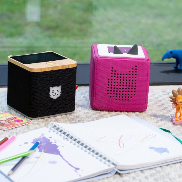 Eine schwarze Tigerbox Touch steht neben einer pinkfarbenen Toniebox, davor liegt ein Ringbuch mit Bunstiften.