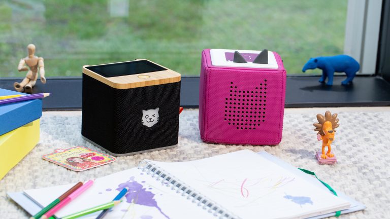 Eine schwarze Tigerbox Touch steht neben einer pinkfarbenen Toniebox, davor liegt ein Ringbuch mit Bunstiften.