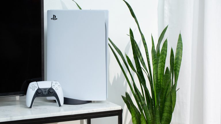 Eine PS5 steht auf einem Marmortisch neben einem Fernseher. Davor lehnt der Dualsense-Controller.