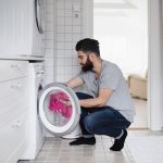 In einem Badezimmer bef&uuml;llt ein Mann die Waschmaschine, &uuml;ber der Waschmaschine steht ein W&auml;schetrockner