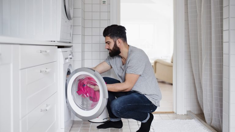 In einem Badezimmer befüllt ein Mann die Waschmaschine, über der Waschmaschine steht ein Wäschetrockner