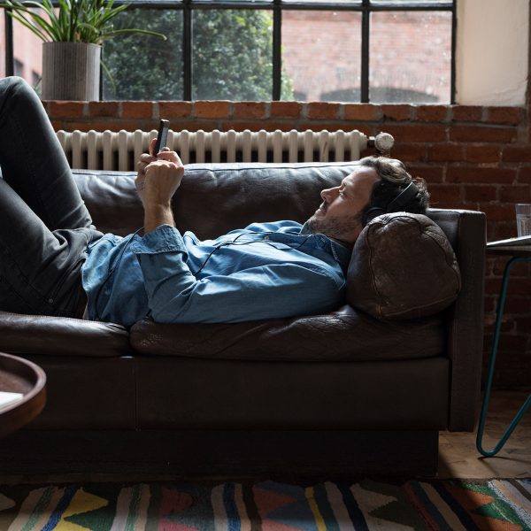 Eine Person liegt auf dem Sofa und h&ouml;rt mit Kopfh&ouml;rern Musik vom Smartphone.