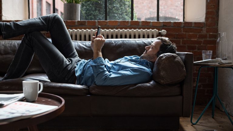 Eine Person liegt auf dem Sofa und hört mit Kopfhörern Musik vom Smartphone.
