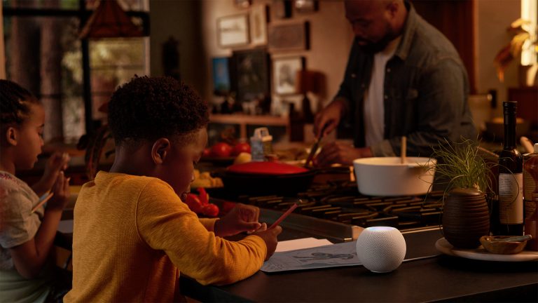 Eine Person und zwei Kinder sitzen an einem Küchentisch. Darauf steht ein Apple HomePod mini.