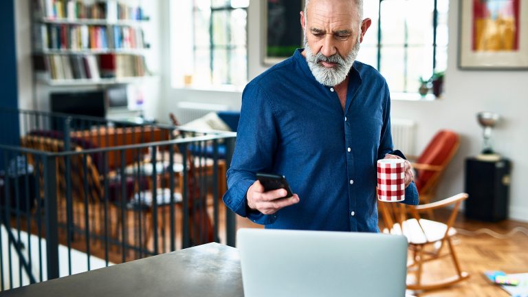 Eine Person steht mit einer Tasse in der Hand vor einem Laptop. In der Hand hält sie ein iPhone.