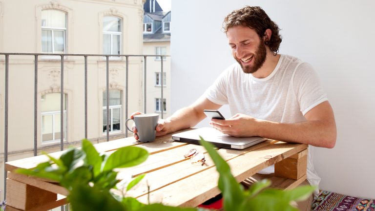 Eine Person sitzt vor einem zugeklappten Laptop und hat ein Smartphone in der Hand.