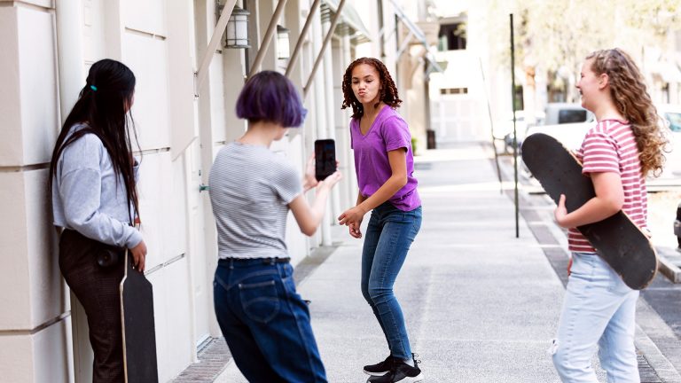 Eine Person steht auf einem Skateboard, eine weitere Person richtet ihr Smartphone auf sie und nimmt ein Video auf.