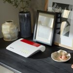 Eine Fritzbox 7590 AX steht auf einer schwarzen Kommode neben Deko-Artikeln.