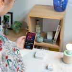 Eine Person steht vor einem Tisch, auf dem ein Bewegungsmelder von Philips Hue, die Philips Hue Bridge und eine Lampe stehen. Sie schaut auf ihr Smartphone, das den Startbildschirm der Philips Hue App zeigt.