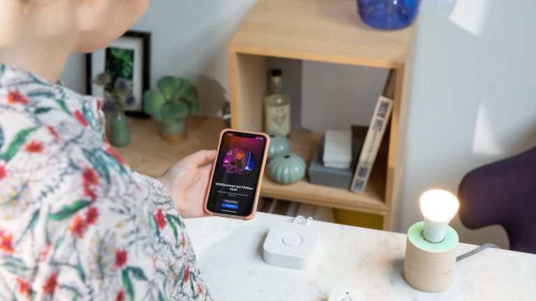 Eine Person steht vor einem Tisch, auf dem ein Bewegungsmelder von Philips Hue, die Philips Hue Bridge und eine Lampe stehen. Sie schaut auf ihr Smartphone, das den Startbildschirm der Philips Hue App zeigt.
