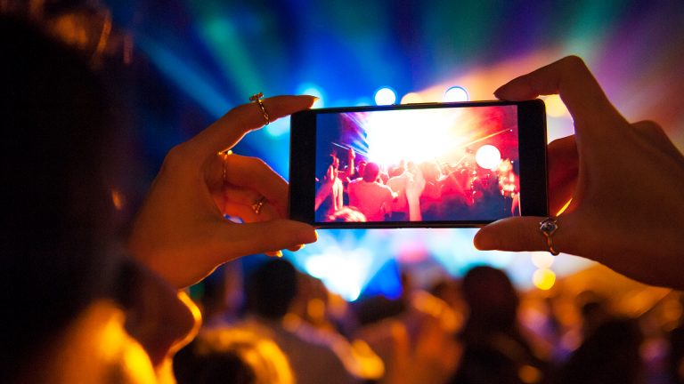 Ein Smartphone wird bei einem Konzert in die Höhe gehalten. Auf dem Bildschirm ist das Publikum zu sehen.