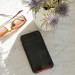 Ein Smartphone liegt auf einem Marmortisch im Schatten. Daneben liegt eine Sonnenbrille.