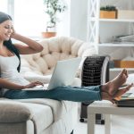 Eine Person sitzt mit einem Laptop auf dem Sofa. Im Hintergrund ist ein Luftk&uuml;hler zu sehen, der auf sie gerichtet ist.