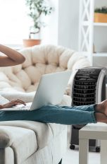 Eine Person sitzt mit einem Laptop auf dem Sofa. Im Hintergrund ist ein Luftkühler zu sehen, der auf sie gerichtet ist.