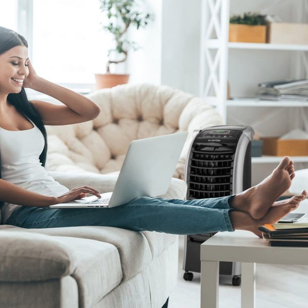 Eine Person sitzt mit einem Laptop auf dem Sofa. Im Hintergrund ist ein Luftk&uuml;hler zu sehen, der auf sie gerichtet ist.
