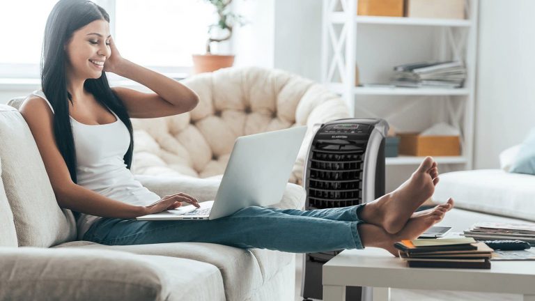 Eine Person sitzt mit einem Laptop auf dem Sofa. Im Hintergrund ist ein Luftkühler zu sehen, der auf sie gerichtet ist.