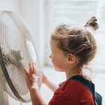 Ein Kind steht vor einem eingeschalteten Ventilator, seine Haare wehen im Wind.