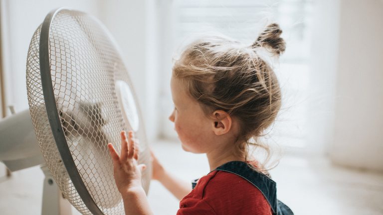 Ein Kind steht vor einem eingeschalteten Ventilator, seine Haare wehen im Wind.