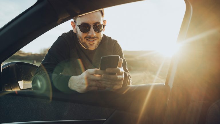 Ein Mann mit Sonnenbrille lehnt sich in ein Fahrzeug. Dabei schaut er auf sein Mobiltelefon.