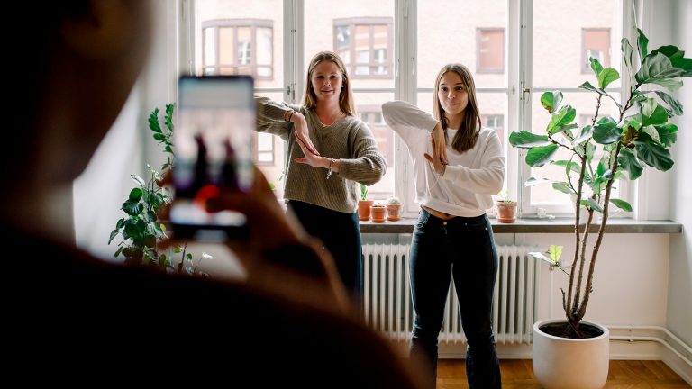 Zwei junge Frauen tanzen eine Choreografie für ein Video. Im Vordergrund ist unscharf ein Smartphone zu erkennen, das die beiden filmt.