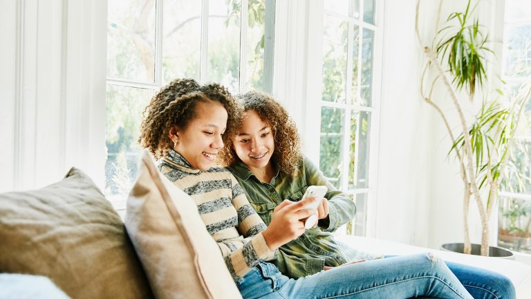 Zwei junge Frauen sitzen auf einem Sofa und schaue gemeinsam auf ein Smartphone.