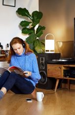 Eine Frau sitzt auf dem Fußboden und liest in einem Magazin, hinter ihr sind ein Sideboard mit TV-Gerät und Lautsprechern rechts und links zu sehen.