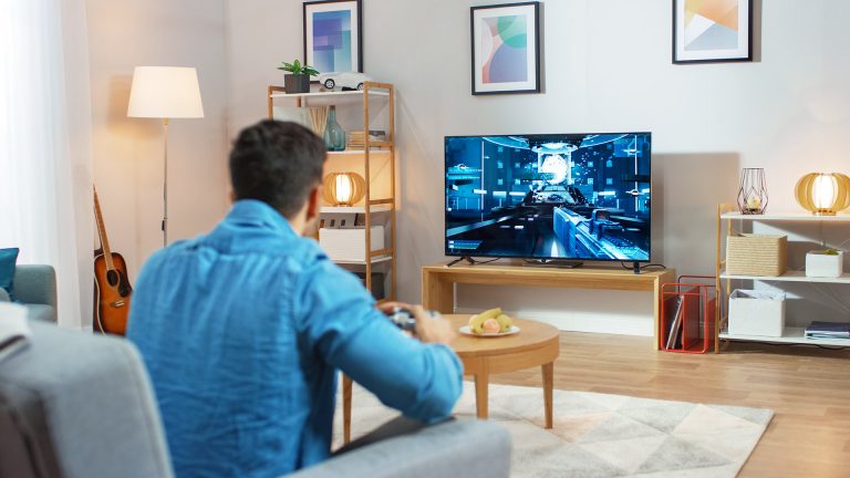 Ein Mann sitzt mit einem Controller auf dem Sofa und spielt ein Spiel auf dem Fernseher.