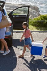Zwei Personen beladen ein Auto. Ihre beiden Kinder tragen zusammen eine Kühlbox zum Wagen.