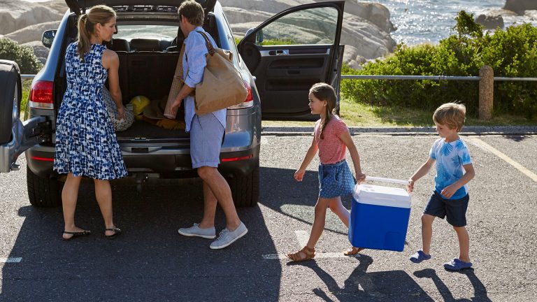 Zwei Personen beladen ein Auto. Ihre beiden Kinder tragen zusammen eine Kühlbox zum Wagen.