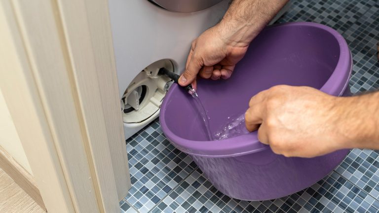 Eine Person lässt über den Ablass-Schlauch einer Waschmaschine überschüssiges Wasser in einen Eimer ab.