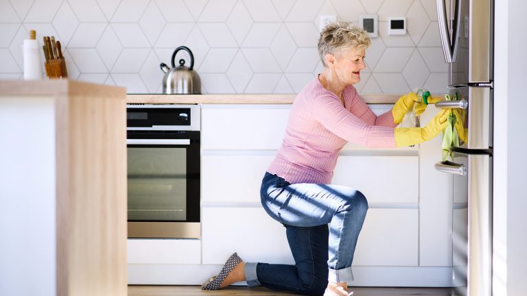 Eine Person kniet vor einem Kühlschrank und macht die Fronten des Geräts mit Putzmittel sauber.