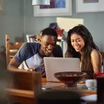 Zwei Personen sitzen vor einem Notebook, blicken auf den Bildschirm und lachen dabei.