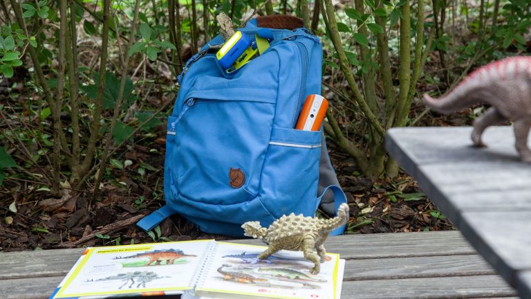 Auf einer Holzbank steht ein blauer Rucksack, davor liegt ein aufgeschlagenes Buch, auf dem ein Spielzeugdinosaurier steht.