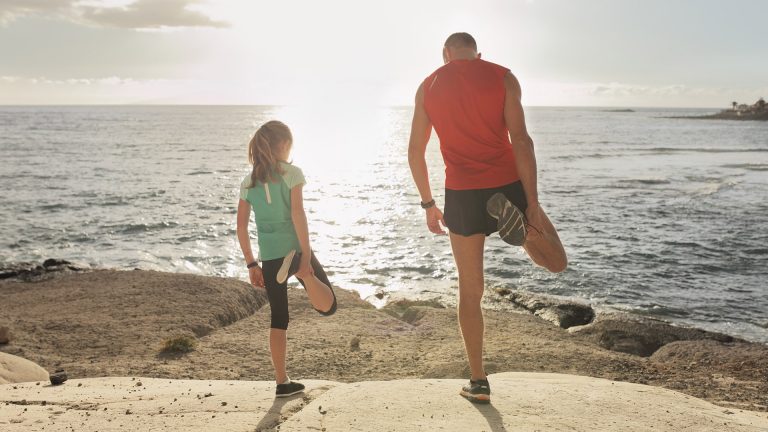 Ein Vater steht mit seiner Tochter auf den Felsen am Meer und macht Dehnungsübungen.