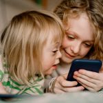 Zwei Kinder liegen auf dem Bett und schauen auf ein Smartphone.