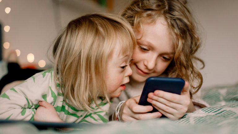 Zwei Kinder liegen auf dem Bett und schauen auf ein Smartphone.