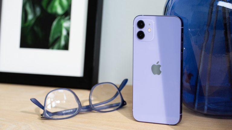 Ein iPhone 12 steht auf einem Holztisch, daneben liegt eine Brille und etwas Deko steht im Hintergrund.
