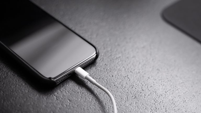 Ein iPhone liegt ausgeschaltet auf einer schwarzen Unterlage. Es ist an ein Kabel angeschlossen.