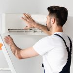 Eine Person auf einer Leiter schraubt die Abdeckung einer Split-Klimaanlage auf.