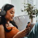 Eine junge Frau sitzt auf dem Sofa, schaut auf ihr Smartphone und h&ouml;rt &uuml;ber einen Kopfh&ouml;rer Musik.