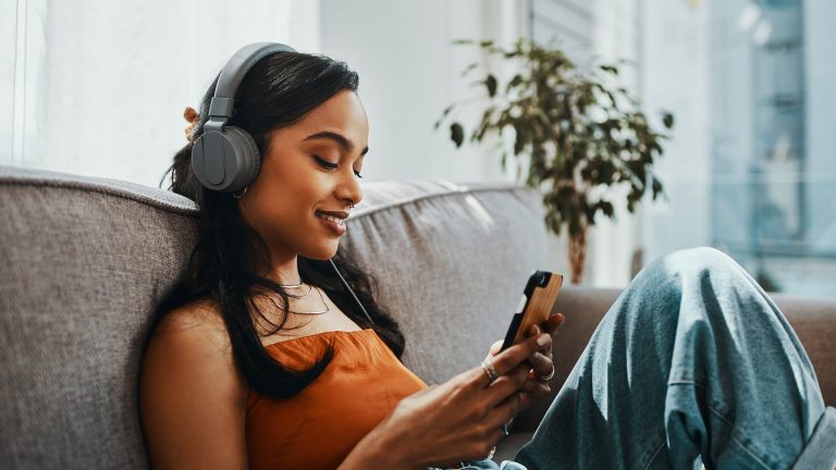 Eine junge Frau sitzt auf dem Sofa, schaut auf ihr Smartphone und hört über einen Kopfhörer Musik.