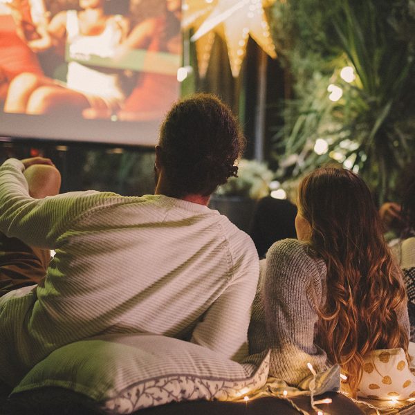Ein Paar liegt auf Kissen und Decken vor einer Leinwand und schaut einen Film im eigenen Garten.