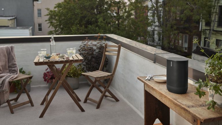Auf einem Balkon steht ein Bluetooth-Lautsprecher auf einem Tisch, im Hintergrund ein kleiner gedeckter Tisch f&uuml;r Zwei.